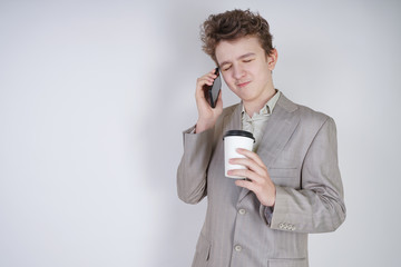 caucasian teenager in a gray business suit stands with a mobile phone and a paper Cup of coffee on a white background in the Studio
