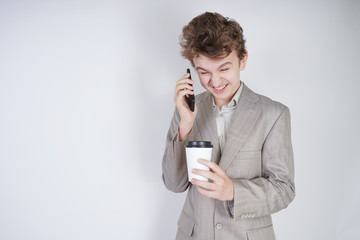 caucasian teenager in a gray business suit stands with a mobile phone and a paper Cup of coffee on a white background in the Studio