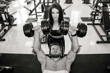 Top view of a beautiful woman in sportswear helping strong man to lift dumbbells. Black and white photo of a beautiful couple in sports gym exercising with dumbbells.