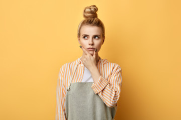 Housewife, chef, cook or baker in gray apron, white t-shirt, striped shirt ,standing with puzzled expression. isolated on yellow background.woman making up a business plan.idea concept