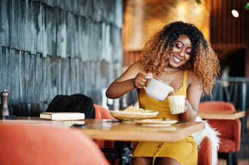 Glamour african american woman in yellow dress sitting on restaurant and pours tea from the teapot.