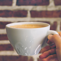 Hand holding cup of cappuccino coffee or orange thai tea on red brick wall background in cafe, toned. Close-up, copy space, square format
