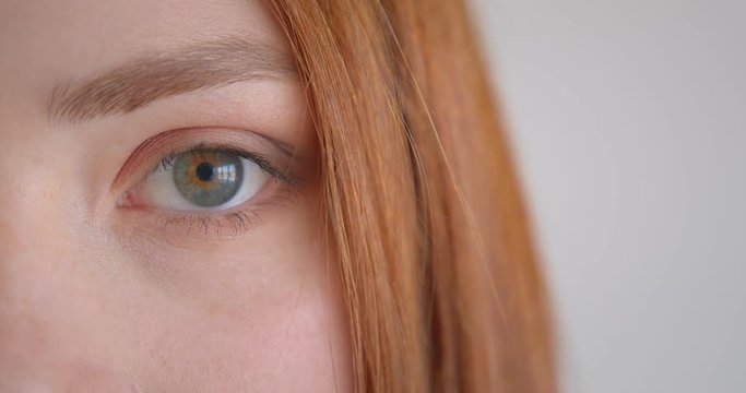 Close-up one eye portrait of beautiful ginger model blinking watching calmly into camera on white background.