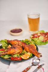 Grilled chicken legs roasted on the grill on dark plate with tomato sauce in a bowl and lettuce leaves, glass mug of beer