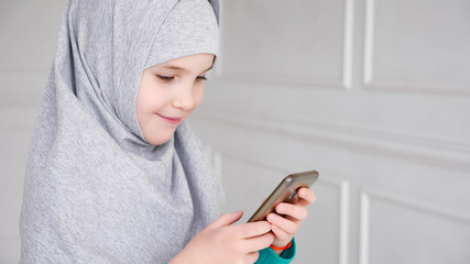 Portrait of young muslim teen girl in grey hijab is playing smartphone, side view.