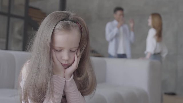Little sad girl sitting on the sofa in the foreground with her head on hands while african ameican father and caucasian mother emotionally talking on the background. Lonely child, family relationship