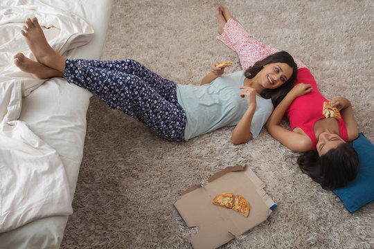 Top view of two smiling young girls lying on the floor in their house eating pizza and talking	
