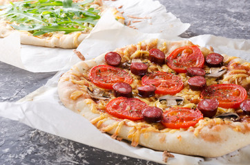 Closeup of tasty hot pizza with tomatoes, cheese, slices of grilled sausages, mushrooms and second one with arugular, different kinds of cheese.Pizza on the baking paper