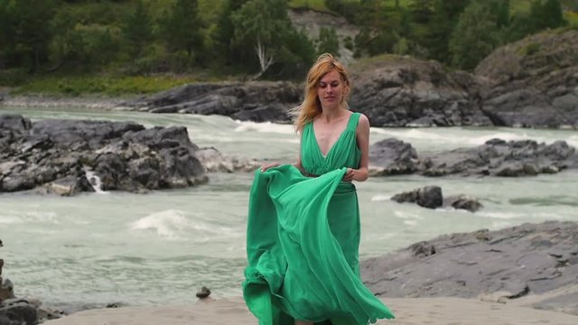 young woman in a long flowing dress on the background of the river and mountains. coloring women goes in a green dress on a background of a beautiful landscape. a woman walks beside a river. soft