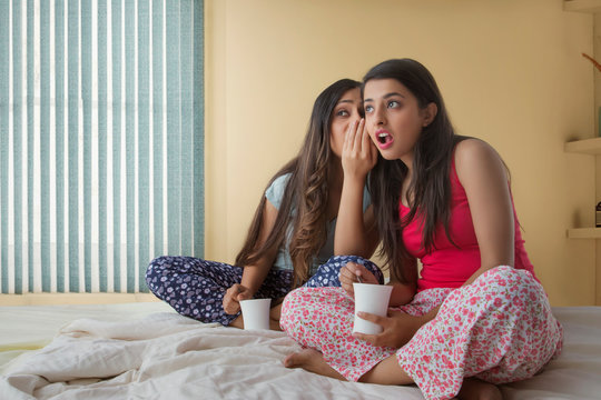 Two young girls sitting on bed at home holding coffee mugs and gossiping	