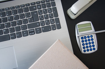 Closeup of laptop, stapler, calculator, pen and notepad on dark background.