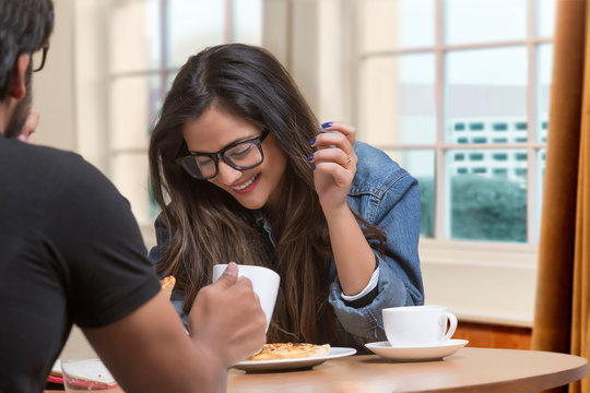 Smiling young girl in spectacles talking to friend sitting at restaurant having pizza and coffee	