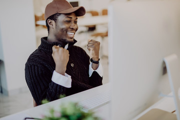 Fototapeta premium Happy african-american businessman looking at laptop excited by good news online. Black man winner sitting at office desk achieved goal raising hands celebrating business success win result