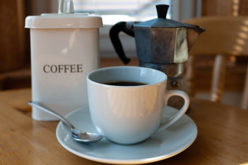 Coffee in a white cup and saucer with silver spoon, a tin of coffee beans, coffee beans and a coffee maker on a kitchen table in bright sunlight