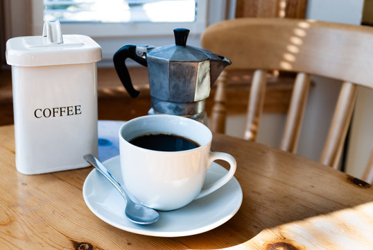 Coffee in a white cup and saucer with silver spoon, a tin of coffee beans, coffee beans and a coffee maker on a kitchen table in bright sunlight
