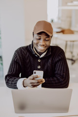 Working with smile. Young African man use phone and working on laptop while sitting at his working place