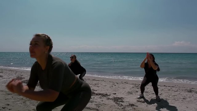 Sports girls on the beach by the sea engaged in group training. In the foreground, the coach shows the exercise. They do jump squats.