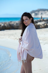 Young girl is on the beach and standing, posing to the camera. Beautiful girl, wrapped in pink white striped beach towel, sunny and windy weather. Background of blue sky, sea and beach - Image