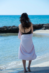 Fototapeta na wymiar Young girl is on the beach and standing, posing to the camera. Beautiful girl, wrapped in pink white striped beach towel, sunny and windy weather. Background of blue sky, sea and beach - Image