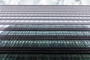 The Facade Of The Office Building Shot From The Bottom Up With A Horizontal Strip Of Gray Sky With Clouds. The Windows Of The Business Center Reflects The Adjacent Building