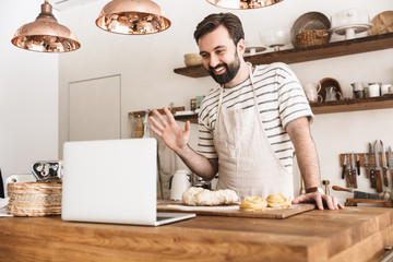 Fototapeta na wymiar Portrait of smiling brunette man using laptop while cooking in kitchen at home