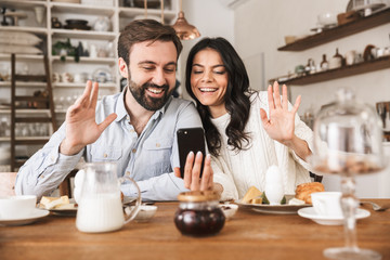 Portrait of smiling european couple using smartphone while having breakfast in kitchen at home