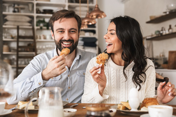 Image of pretty couple eating together at table while having breakfast in kitchen at home