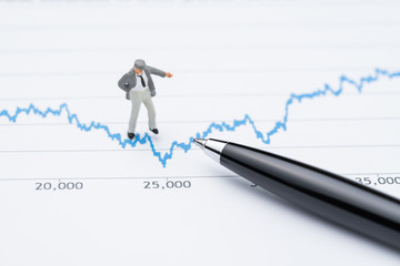 Marketing analysis or stock investment volatility concept, miniature professional businessman standing and looking, review on up and down blue stock market line graph data report with pen