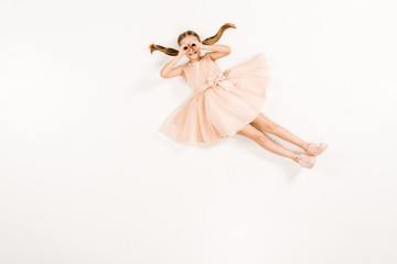 cheerful kid in dress with hands near eyes while lying on white