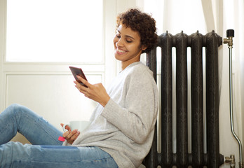 young african american woman sitting on floor next to radiator and looking at mobile phone
