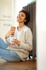 young african american woman sitting on floor against radiator heater with cup of tea and cellphone