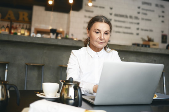 Picture of attractive middle aged businesswoman in formal wear browsing website for job search and staff recruitment on generic portable computer during break at coffee shop, having concentrated look