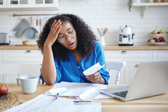 People, modern technology, finance and housework concept. Picture of tired young dark skinned woman in eyeglasses doing paperwork at home, having frustrated look, shocked with amount of debt