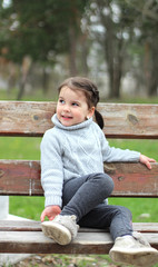 beautiful little girl with ponytails and in a sweater sitting on a bench in the park and smiling