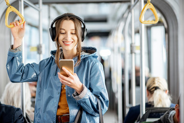 Young woman passenger standing with headphones and smartphone while moving in the modern tram, enjoying trip at the public transport - obrazy, fototapety, plakaty