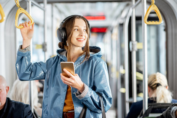 Young woman passenger standing with headphones and smartphone while moving in the modern tram, enjoying trip at the public transport - obrazy, fototapety, plakaty