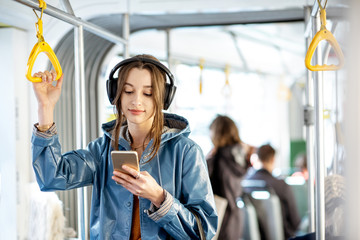 Young woman passenger standing with headphones and smartphone while moving in the modern tram