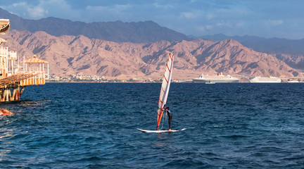a child windsurfer sailing into the eilat marina in israel with the port of akaba jordan in the background