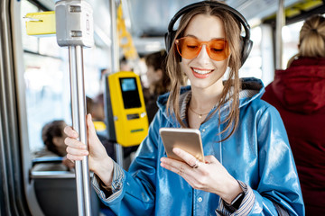 Young stylish woman using public transport, standing with headphones and smartphone while moving in...