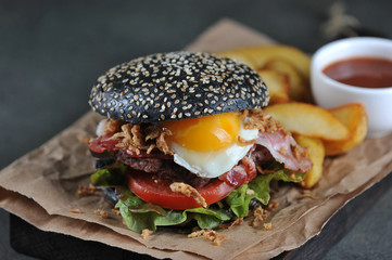 Black burger with fried egg. In the burger filling bacon and onion chips. A rustic potato next to a burger. Dark background. Close-up. Close-up.