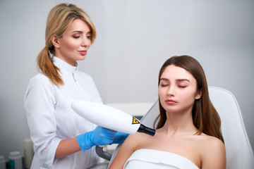 Fototapeta premium Laser mole removal on a woman's chest in a beauty salon. Hardware cosmetology. Pretty blonde beautician doctor removing birthmark or nevus with laser beam gun in beauty salon.