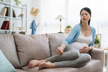 Obraz na płótnie Canvas young asian pregnant woman hands massaging swollen feet while sitting on sofa. illness future mom with cramp in legs hurting putting on couch release painful in living room at home with frowning face