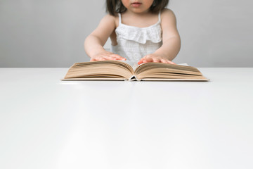 The little girl opened the book. The child is studying the book. A girl is reading a book. The book is on a white table. The girl in white overalls. Girl with a short haircut