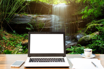 Mockup image of laptop with blank white screen,smart phone and document on wooden table at Waterfall in Tropical Rainforest Landscape,Leisure and travel concept.