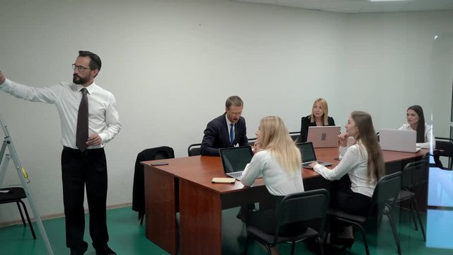 A businessman discussing a flip chart on a white board in a meeting room to working team while standing in the office.