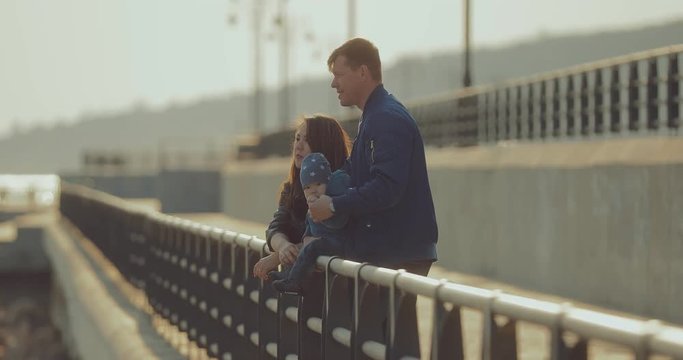 Husband and wife stand at the parapet and hold their little son. Baby smiles, parents show off. Happy family walk.