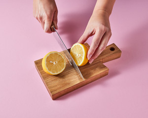 Woman's hands cut a lemon on a wooden board with a knife around a pink background with copy space. Ingredient for summer drink