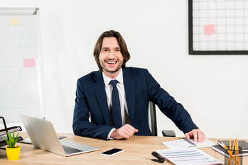 cheerful man in formal wear smiling near laptop and smartphone in office