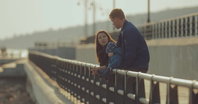 Korean woman and her husband are standing on the riverbank near the parapet and talking. Father holds a little son. International family walks by the river.