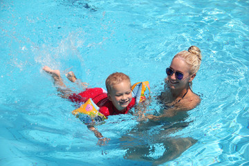 Young woman teaches a little boy to swim in swimming pool on sunny summer day, child in armlets learns having fun with mother in the pool, family vacation and holiday with children concept